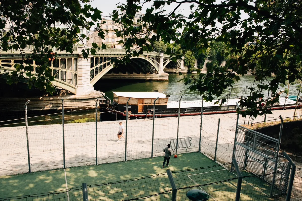 People playing sports near the scenic Seine river with iconic Paris bridge and trees overhead.